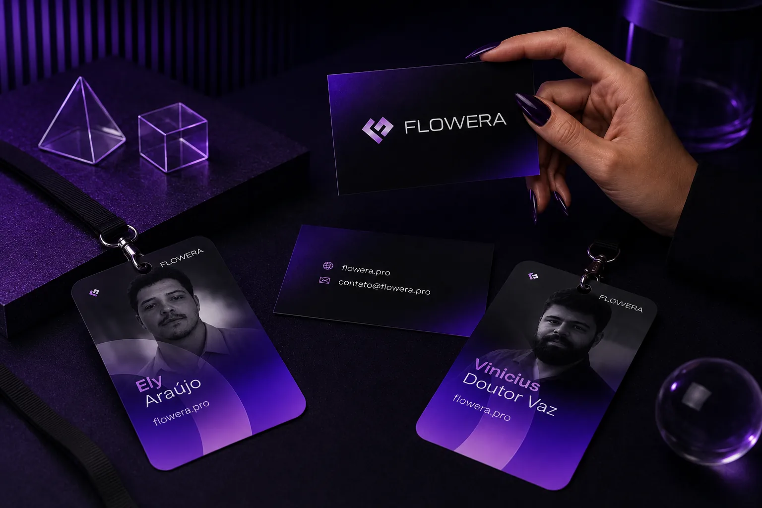 Papelaria da Flowera sobre uma mesa escura: uma mão segura o cartão de visita com a marca, o verso traz flowera.pro e contato@flowera.pro, e dois crachás de cordão mostram os retratos de Ely Araújo e Vinicius Doutor Vaz.